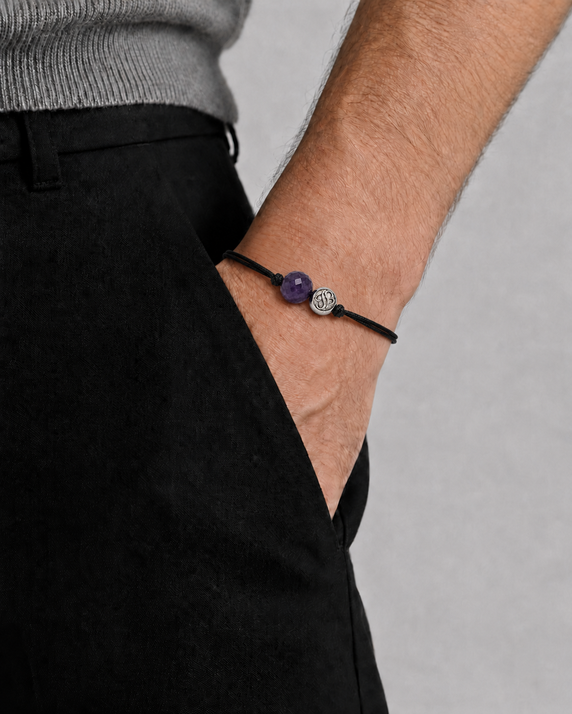 Person wearing a bracelet with a purple bead and silver charm on a plain background